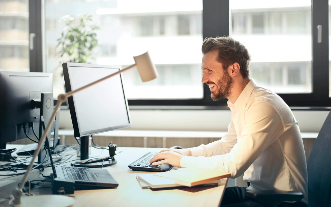 A man in a white shirt is smiling while working at a computer desk in a bright office in Orlando.