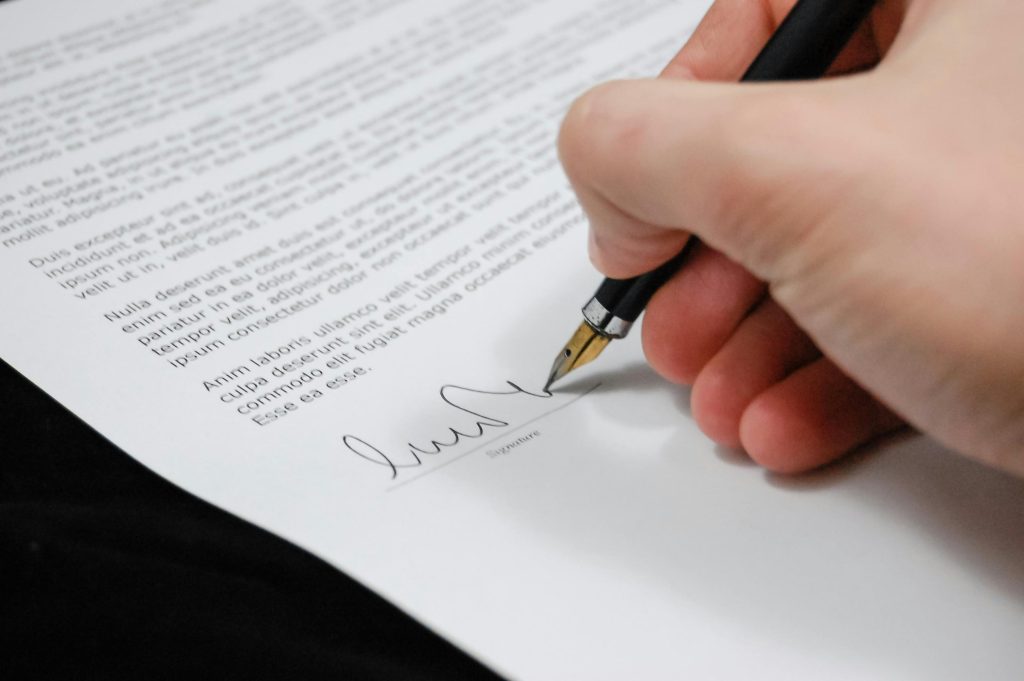 A hand signing a document with a fountain pen in Tampa; the paper contains dense text and a signature line at the bottom.