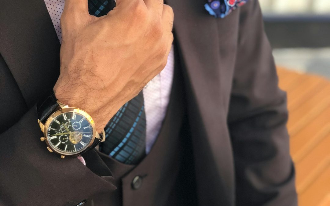 A man is adjusting a blue striped tie, complementing his brown suit and vest. His outfit is accentuated with a gold wristwatch and a blue-patterned pocket square, embodying the classic style often seen in Tampa.